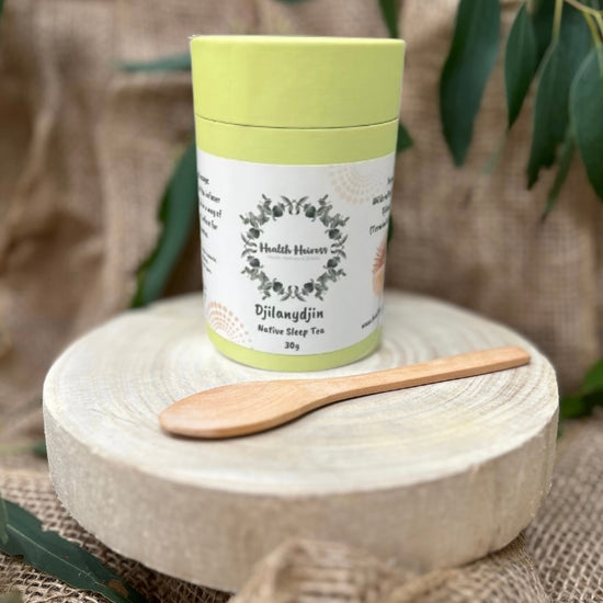 A green cylindrical container labeled 'Djilanydjin Native Bush Tea' with a wooden spoon beside it, placed on a natural fiber mat with leaves in the background.