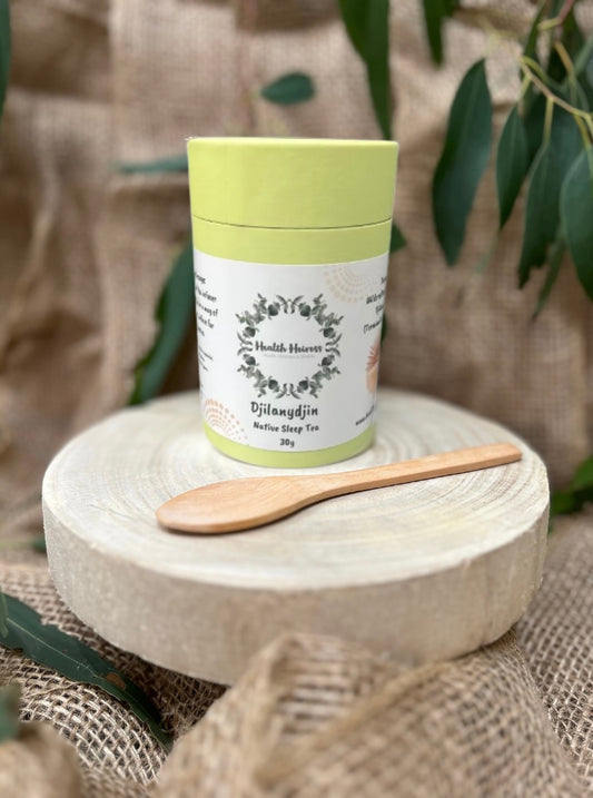 A green cylindrical container labeled 'Djilanydjin Native Bush Tea' with a wooden spoon beside it, placed on a natural fiber mat with leaves in the background.